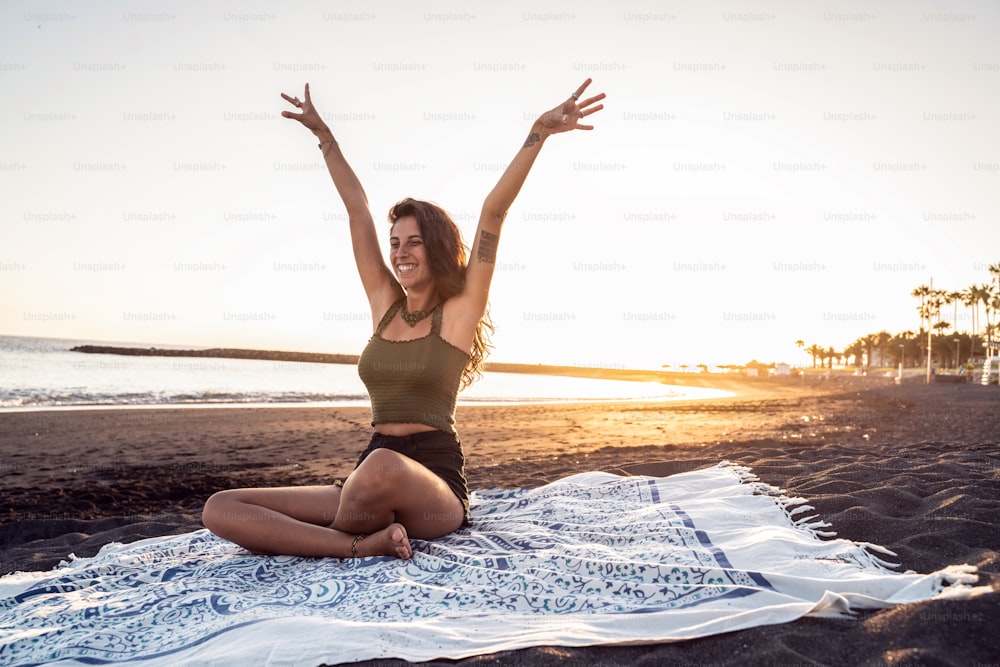 Happy carefree beautiful woman enjoying sunset evening on the sandy beach, having fun, smiling, sitting on the blanket. Summer positive vibes. Real people emotions, lifestyle.