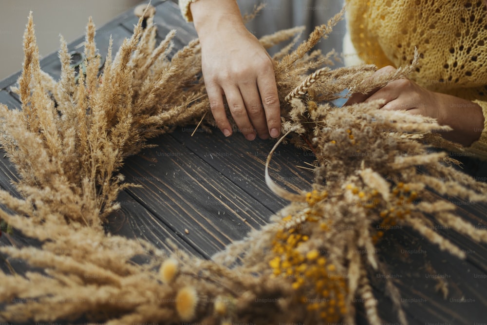Hands making stylish autumn rustic wreath with dry grass, wildflowers and wheat on rustic wooden table in room. Holiday workshop. Florist making boho wreath on dark wood