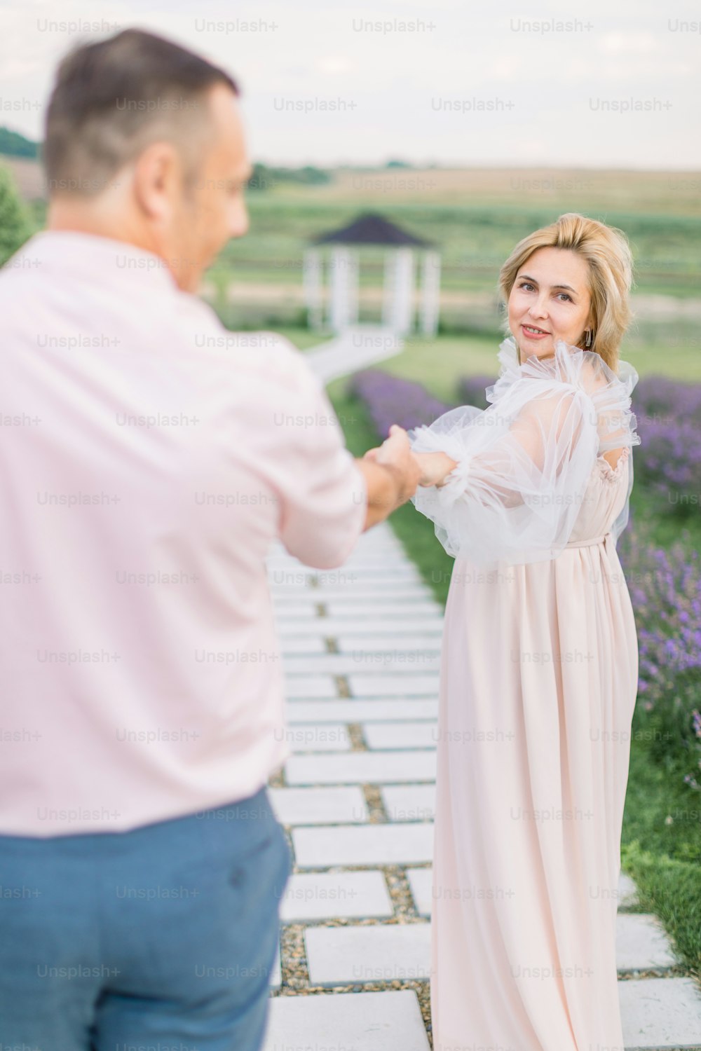 Give me your hand, follow me. Love through the years together. Pretty charming blond mature lady in elegant dress, holding hand and walking with her handsome man outdoors in lavender field with gazebo