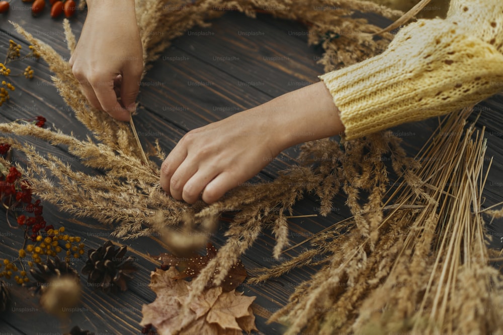 Florist im gelben Pullover bastelt rustikalen Herbstkranz auf dunklem Holztisch. Hände, die trockenes Gras halten und stilvollen Boho-Kranz mit Wildblumen und Kräutern auf rustikalem Holzhintergrund machen.
