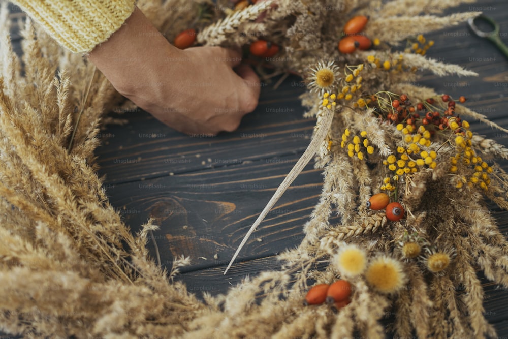 Hands making stylish autumn rustic wreath with dry grass, wildflowers, wheat and berries on rustic wooden table in room. Holiday workshop. Florist making boho wreath on dark wood