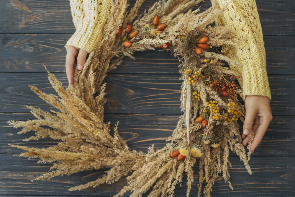 Hände halten stilvollen Herbst-Bauernkranz mit trockenem Gras, Wildblumen, Weizen und Beeren auf rustikalem Holztisch, flach gelegt. Ferienwerkstatt. Florist macht Boho-Kranz auf dunklem Holz