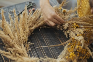 Stilvoller Herbst-Boho-Kranz mit trockenem Gras und Wildblumen auf rustikalem Holztisch. Ferienwerkstatt. Florist im gelben Pullover macht rustikalen Kranz auf dunklem Holz