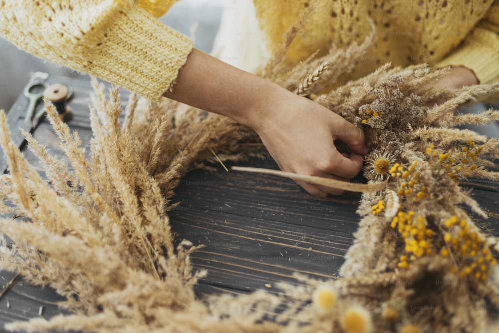 Realizzazione di un'elegante ghirlanda autunnale. Mani che tengono l'erba secca, i fiori di campo e il grano e fanno ghirlanda rustica sul tavolo di legno. Laboratorio per le vacanze autunnali. Fioraio che fa ghirlanda boho su legno scuro