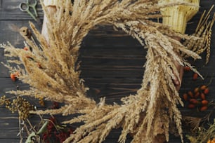 Manos sosteniendo una elegante corona de otoño con hierba seca, flores silvestres, hierbas y bayas sobre fondo de madera rústica. Vista superior. Hacer corona boho rústica de otoño, taller de vacaciones