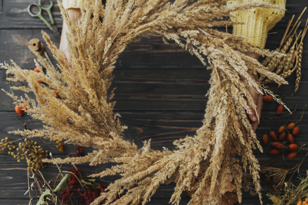 Hands holding stylish autumn wreath with dry grass, wildflowers, herbs and berries on rustic wooden background. Top view. Making rustic autumn boho wreath, holiday workshop