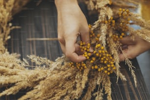 Mains faisant une couronne d’automne rustique élégante avec de l’herbe sèche et des fleurs sauvages de tanaisie sur fond en bois foncé. Atelier de vacances. Fleuriste en pull jaune faisant une couronne bohème sur une table en bois