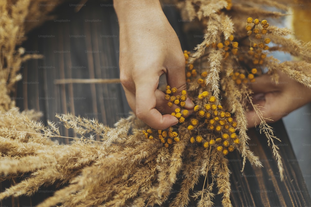 Hands making stylish rustic autumn wreath with dry grass and tansy wildflowers on dark wooden background. Holiday workshop. Florist in yellow sweater making boho wreath on wooden table