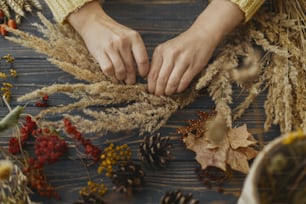 Hände halten trockenes Gras und machen stilvollen Herbst-Boho-Kranz mit Wildblumen und Kräutern auf rustikalem Holztisch. Ferienwerkstatt. Floristin im gelben Pullover macht rustikalen Herbstkranz