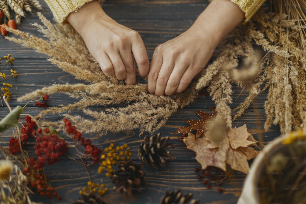 枯れ草を手に、素朴な木製のテーブルの上で野の花やハーブを使ったスタイリッシュな秋の自由奔放に生きるリースを作る手。休日のワークショップ。素朴な秋の花輪を作る黄色のセーターの花屋
