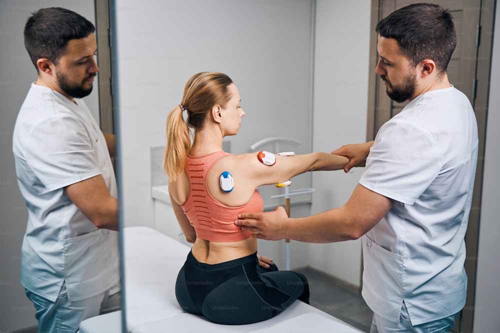 Physical therapy electrodes for myostimulation. Doctor in uniform puts electrostimulator on young woman hand at mirror. Massage with electric stimulator in modern therapy room