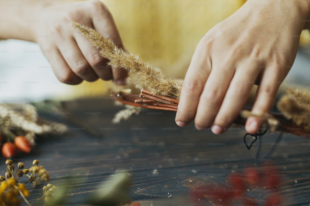 Hands holding dry grass and making stylish autumn boho wreath with wildflowers and herbs on rustic wooden table. Holiday workshop. Florist in yellow sweater making rustic autumn wreath