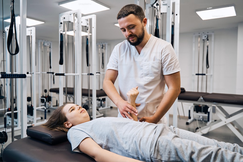 Portrait of physiotherapist massages girls hand in physio room. Professional pediatric therapist treats arm of young female patient at rehab clinic with modern equipment. Caucasian chiropractor man