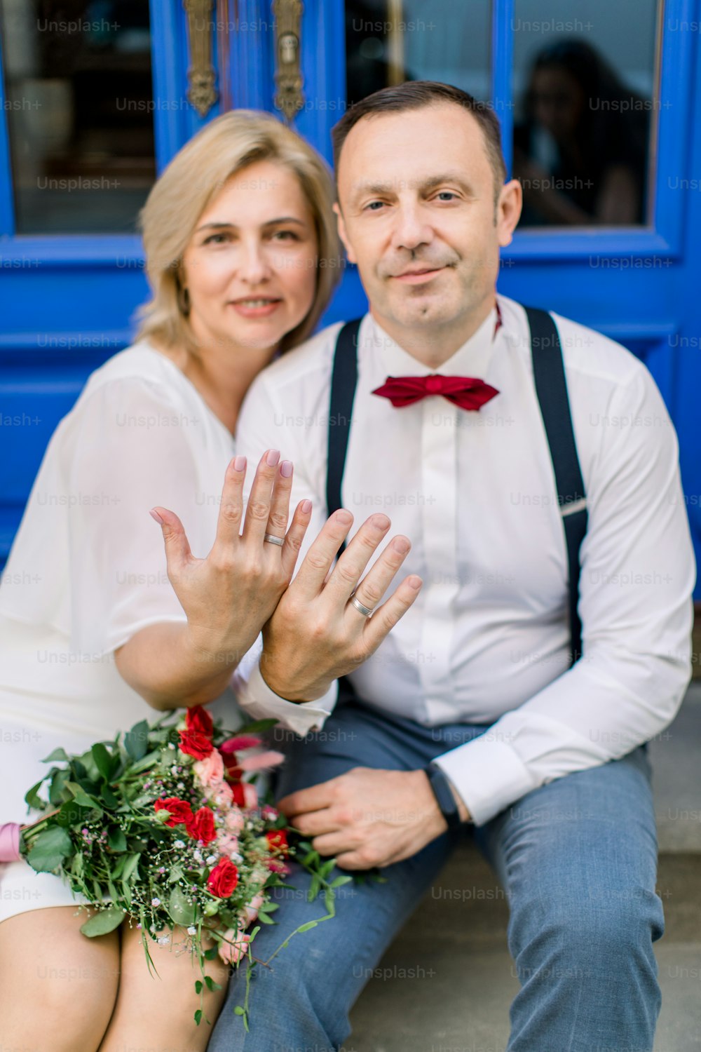 Love through the years. Happy smiling mature couple, sitting in front of blue vintage doors in the city street, showing their hands with the wedding rings. Focus on the rings
