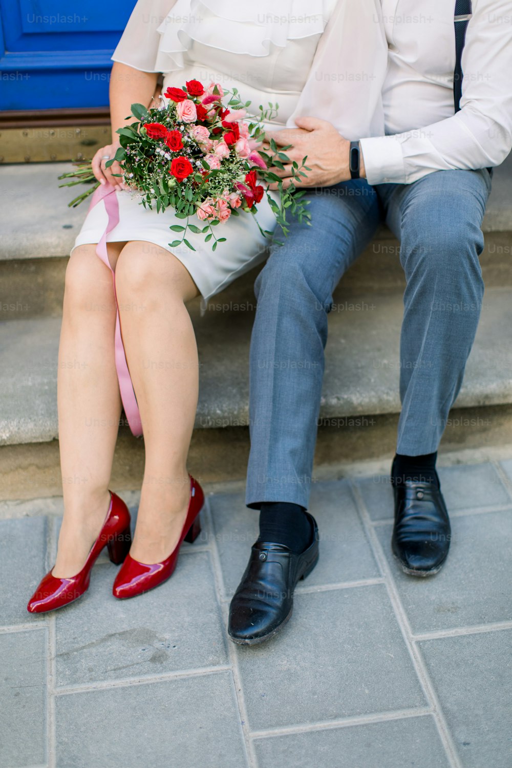 Close up cropped vertical shot of the legs of mature couple, man and woman in elegant suit and dress, sitting on vintage stone stairs with flower bouquet, outdoors on the city street