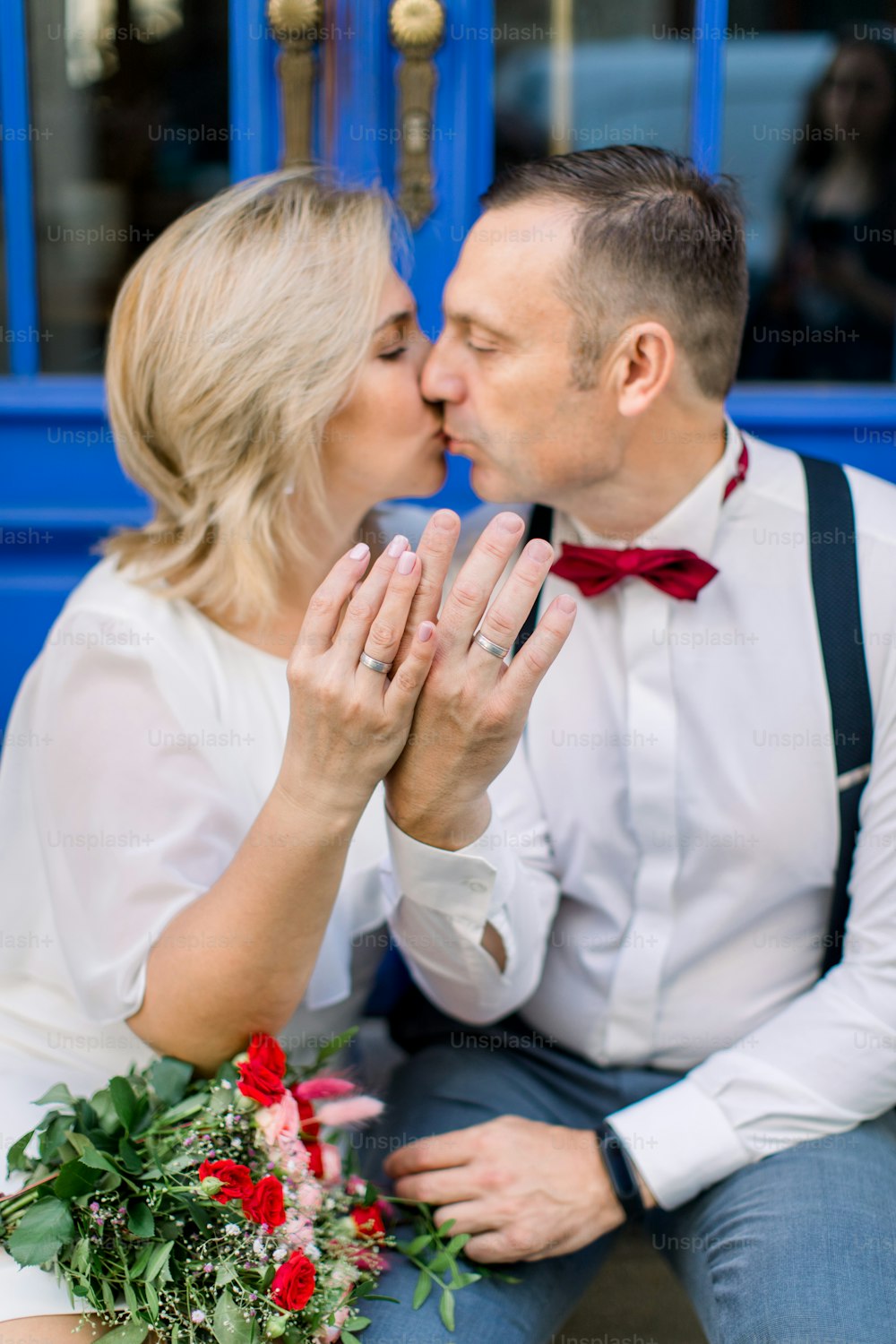 Love through the years. Happy kissing mature couple, sitting in front of blue vintage doors in the city street, showing their hands with the wedding rings. Focus on the rings