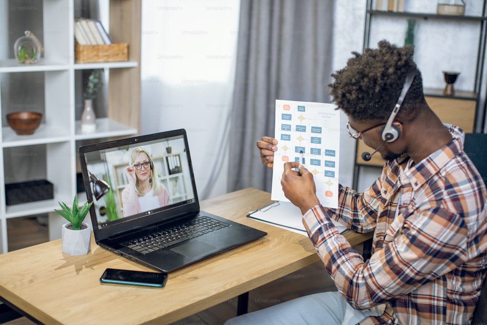 Side view of afro american student passing exams through video call on modern laptop during pandemic time. Caucasian female teacher with blond hair on computer screen.