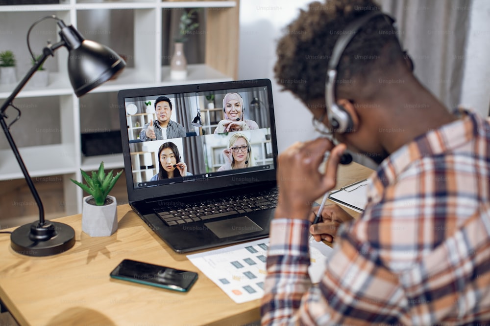Back view of african man in headset talking with coworkers during video call on laptop. International colleagues solving working issues remotely while staying at home.