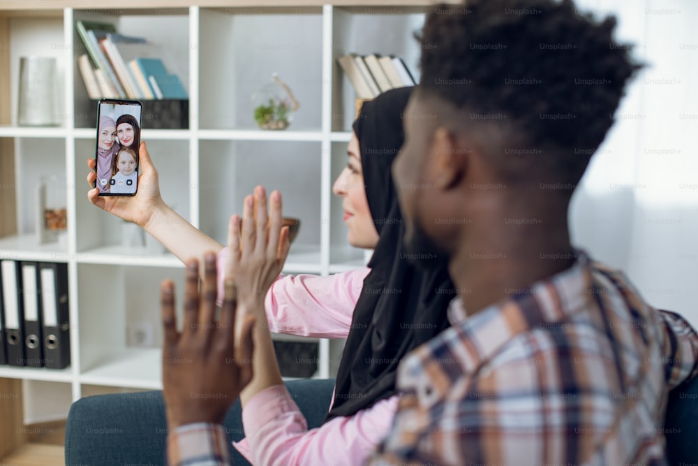 Young international couple sitting at home and talking with family using modern smartphone. African man and muslim woman in hijab having video conversation with relatives.