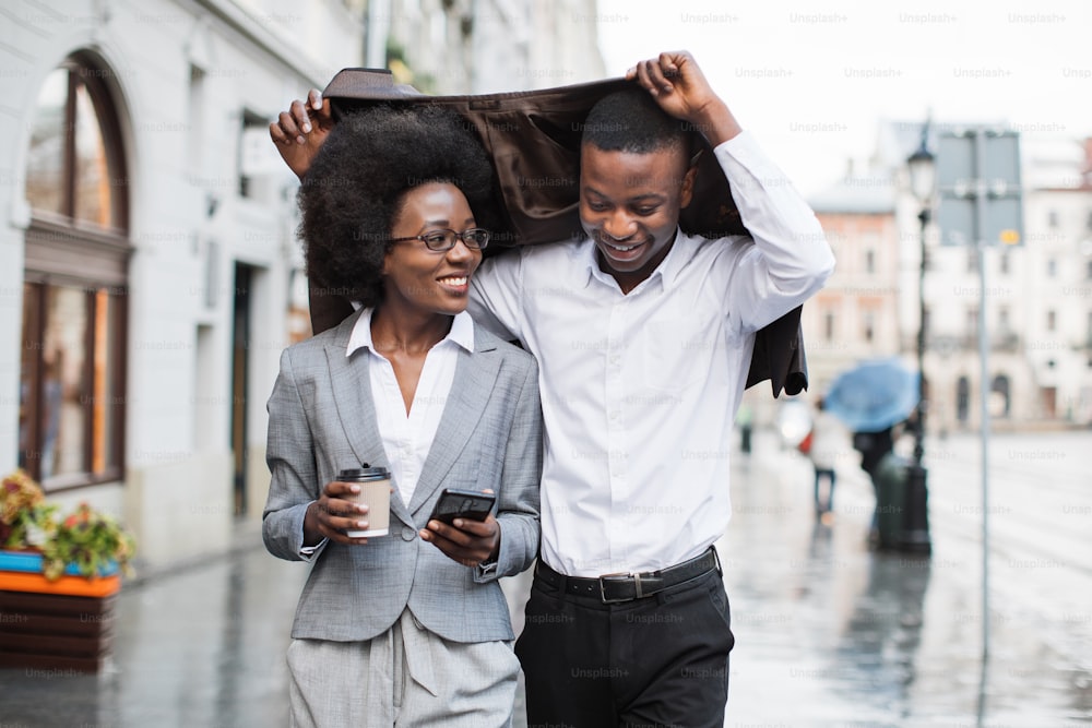 Uomo e donna afroamericani felici che si nascondono dalla pioggia sotto la giacca mentre camminano per la strada della città. Due partner commerciali che vanno alla riunione di lavoro.