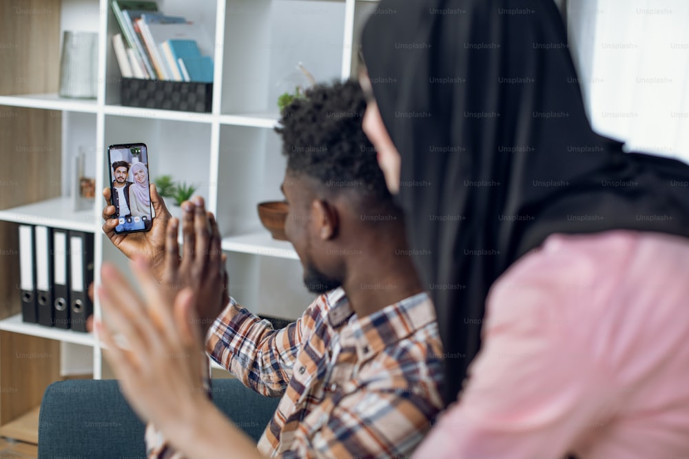 Uomo africano sorridente e donna musulmana che hanno videochiamata su smartphone con i loro parenti. Giovane coppia che guarda lo schermo del cellulare e agita le mani.
