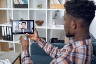 Side view of african male freelancer using digital tablet for video conference with asian coworker. Young man sitting at home and greeting partner on distance.