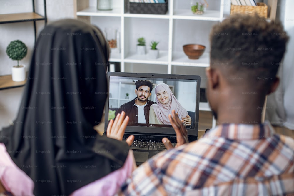 Hombre afroamericano y mujer musulmana con hiyab agitando las manos mientras tienen una videoconferencia con familiares en una computadora portátil. Concepto de pandemia, tecnología y comunicación.