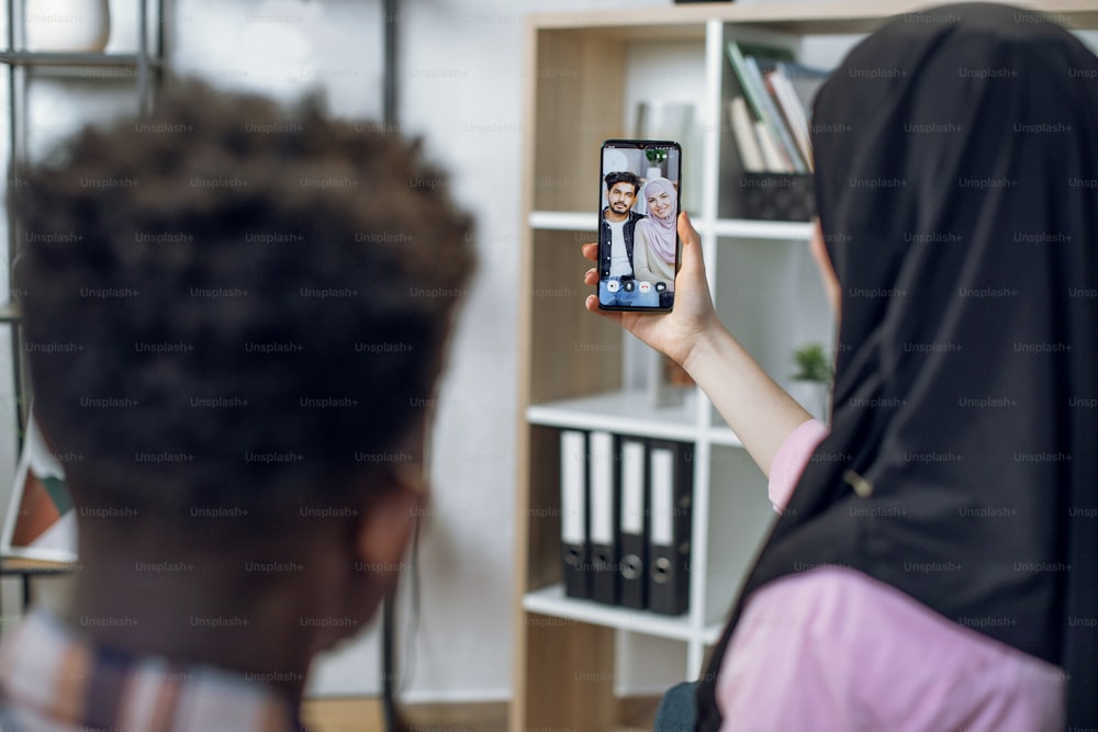 Back view of multiracial couple sitting at home and talking with their muslim friends during video call. People using modern smartphone for remote conversation.