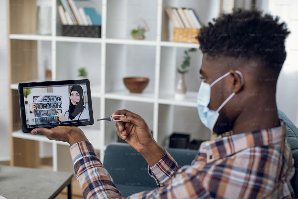 Afro american man in medical mask having video call with muslim woman in hijab on digital tablet. Sick patient with thermometer consulting with competent doctor online.