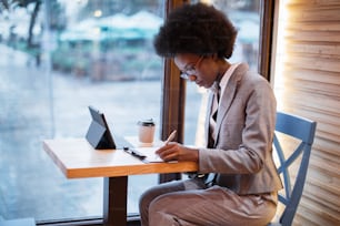 Attractive african woman in eyeglasses and business suit sitting at cafe table, using digital tablet and taking notes. Concept of busy work and technology.