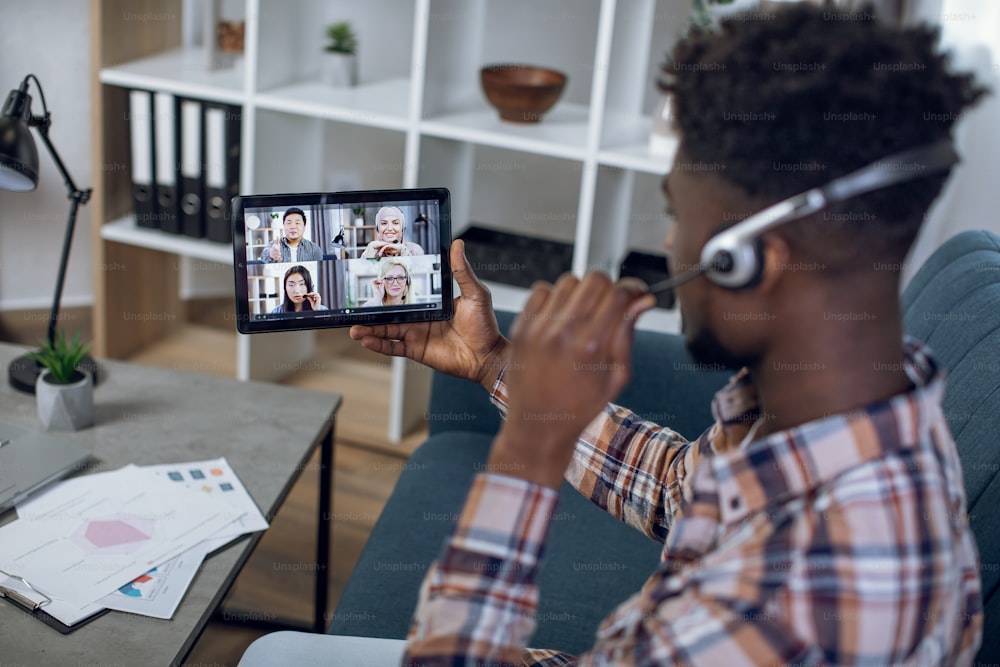 Chico afroamericano con auriculares que tiene una videoconferencia con colegas multirraciales en una tableta digital. Personas diversas que se quedan en casa y cooperan a distancia.