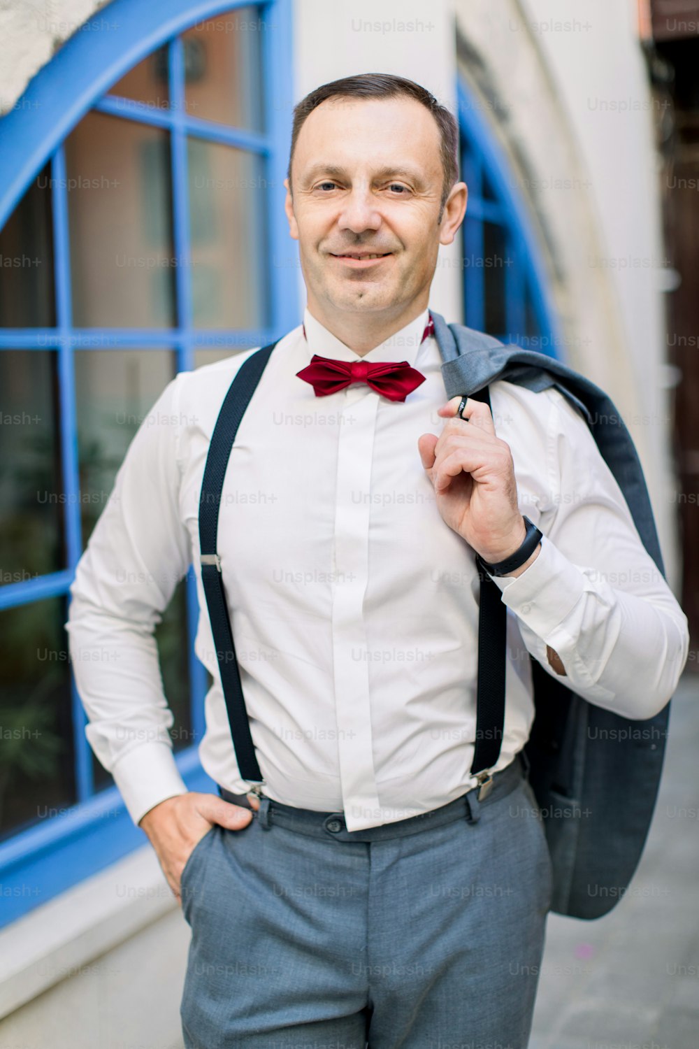 Handsome mature Caucasian man groom in a white shirt with a red bow tie and in suspenders holds a jacket, posing in front of vintage building with big blue window outdoors in the city