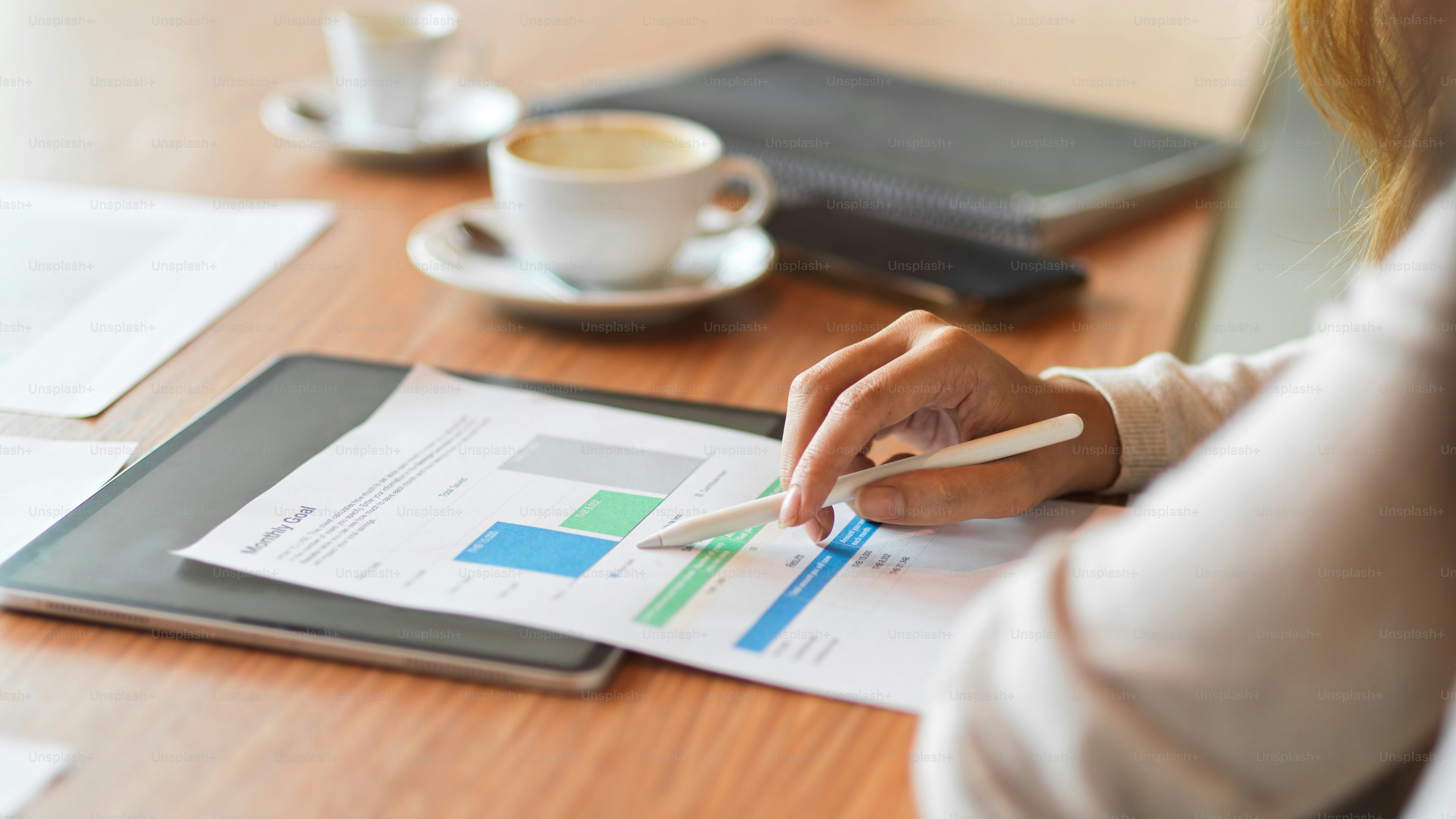 Femme d’affaires lisant un rapport financier tout en tenant un stylet sur le bureau en bois avec une tablette, une tasse à café et des fournitures de bureau