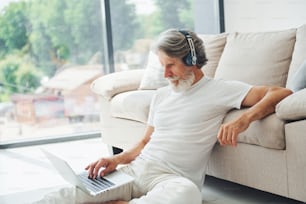 Sits on the ground near sofa with laptop and listens music. Senior stylish modern man with grey hair and beard indoors.