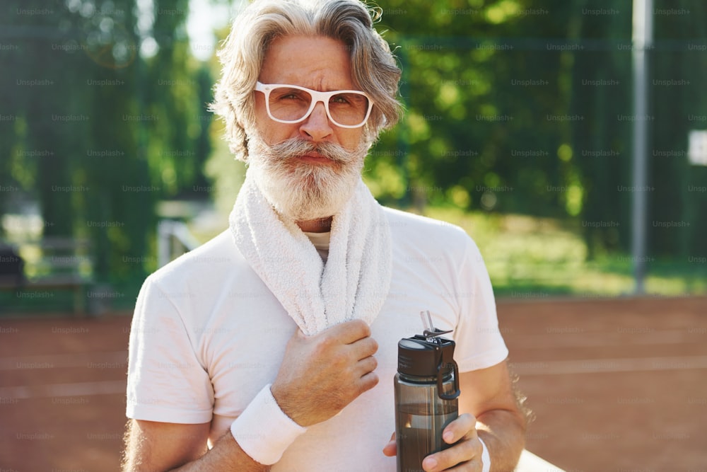 Concezione della moda. Con gli occhiali da sole. Uomo anziano moderno alla moda all'aperto sul campo sportivo durante il giorno.