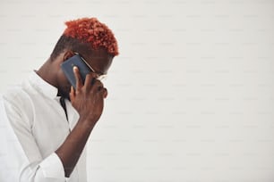 Young african american man in white formal clothes talking by the phone against wall indoors.