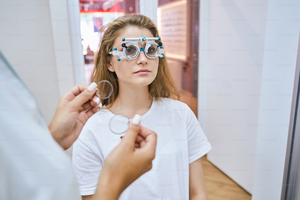 Beautiful young woman using ophthalmic testing device while doctor holding lenses