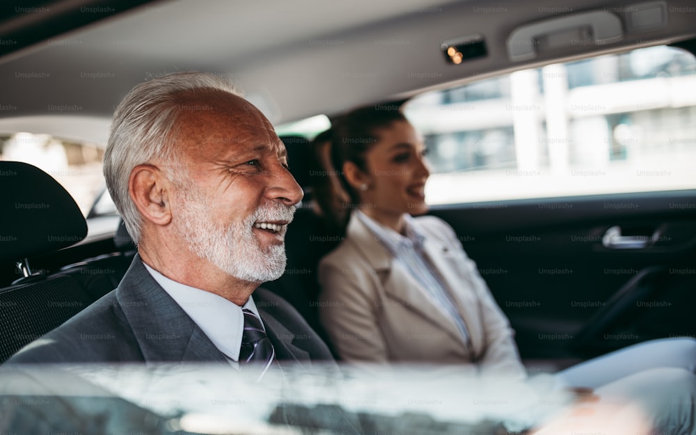 Gut aussehender älterer Geschäftsmann und seine junge Kollegin oder Kollegin sitzen auf dem Rücksitz in einem Luxusauto. Sie unterhalten sich, lächeln und benutzen Laptop und Smartphones. Transport im Unternehmenskonzept.