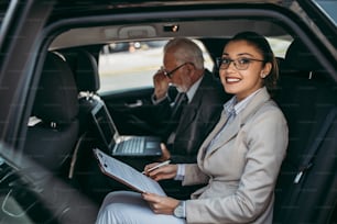 Good looking senior business man and his young woman colleague or coworker sitting on backseat in luxury car. They talking, smiling and using laptop and smart phones. Transportation in corporate business concept.