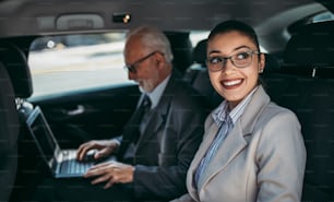 잘 생긴 고위 사업가와 그의 젊은 여성 동료 또는 동료가 고급 자동차의 뒷좌석에 앉아 있습니다. 그들은 이야기하고, 웃고, 노트북과 스마트 폰을 사용합니다. 기업 비즈니스 개념의 교통.