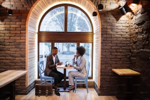 Side view of afro american business people in formal clothes sitting at cafe table drinking coffee, using modern gadgets and communicating. Concept of cooperation and working meeting.