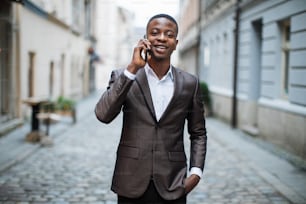 African man in business suit having mobile conversation while walking on city street. Handsome young guy smiling and talking outdoors.