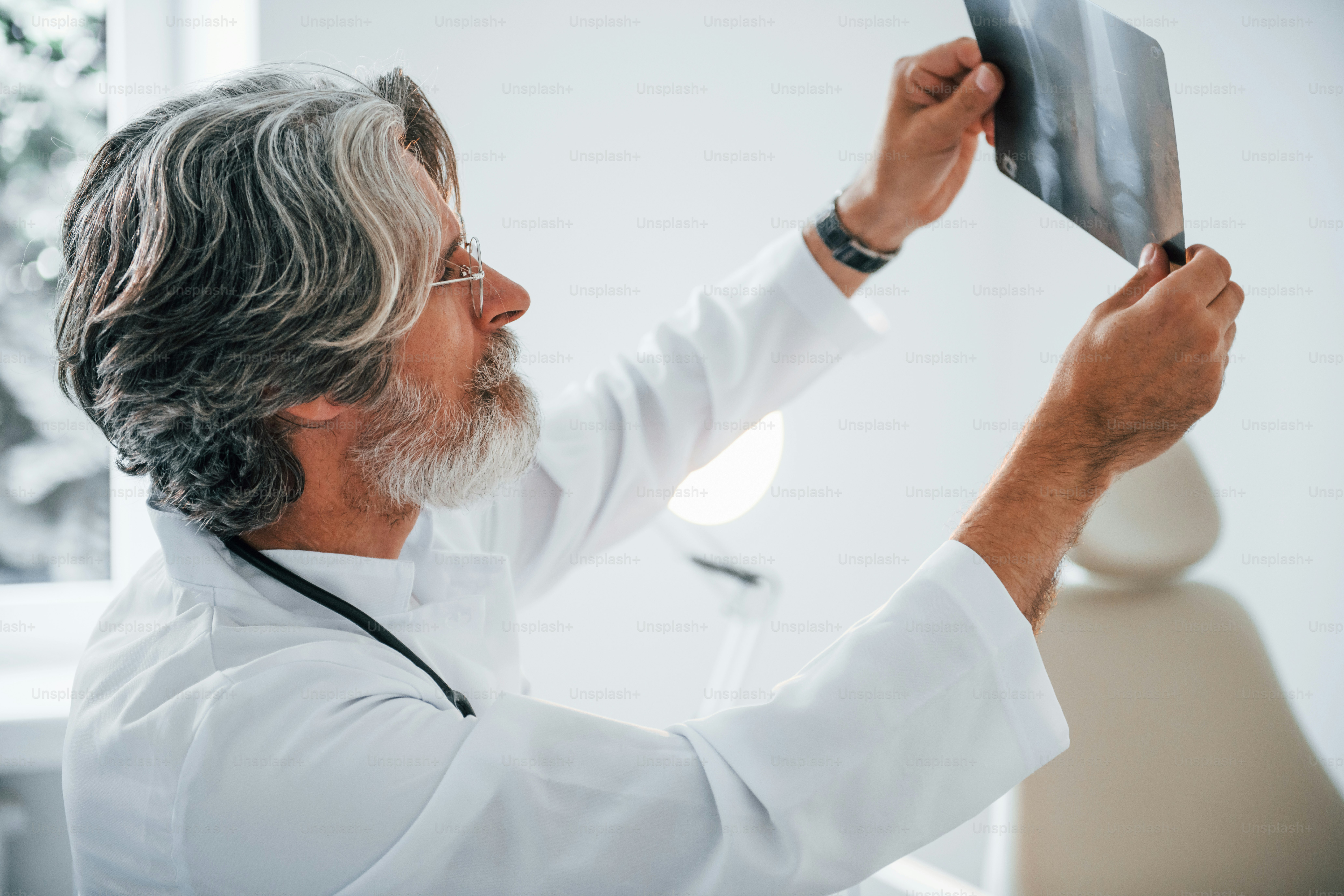 Looks at x-ray. Senior male doctor with grey hair and beard in white coat is indoors in clinic.