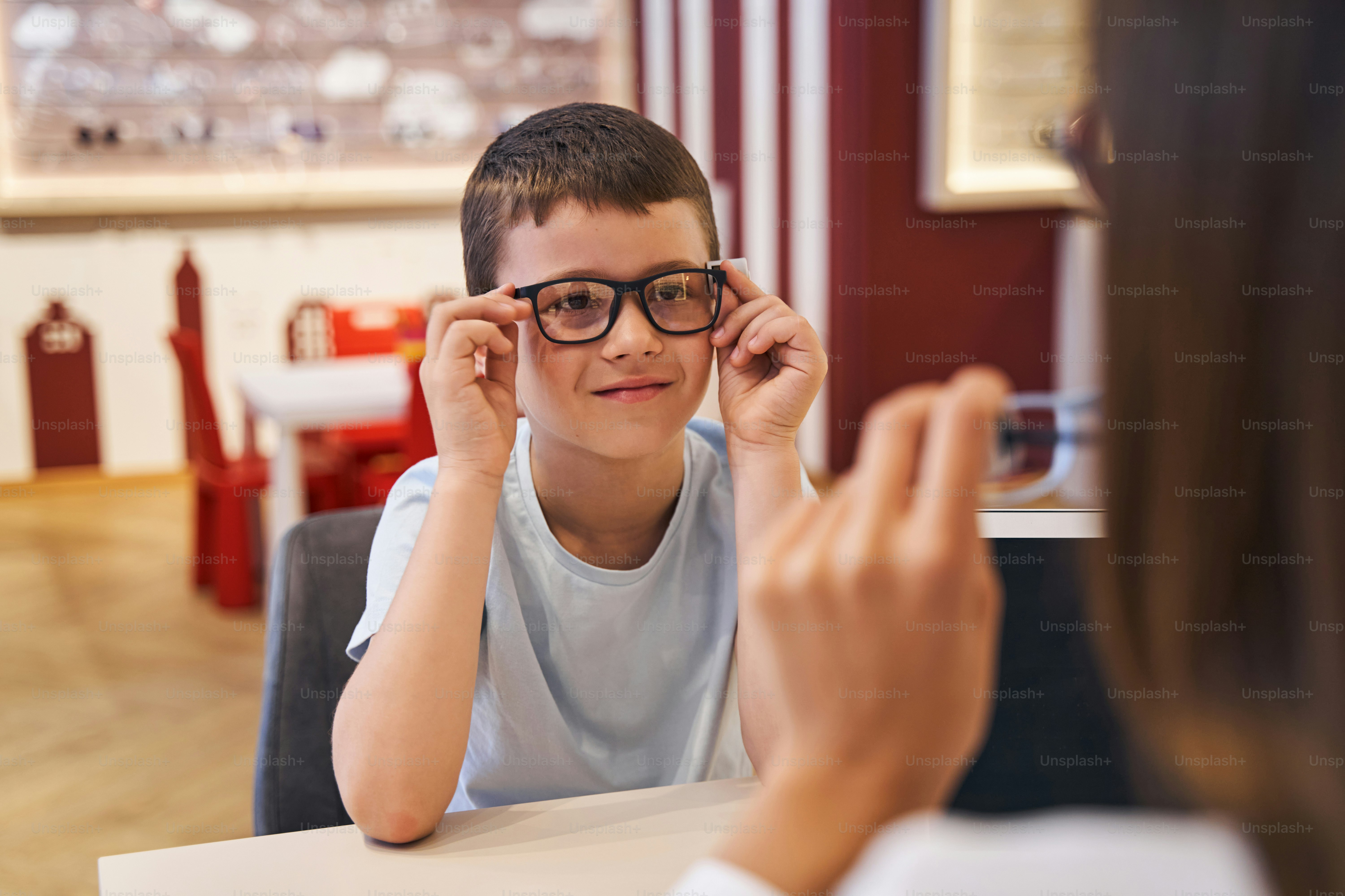 Cute male child wearing eyeglasses and smiling while sitting at the table and looking at optometrist