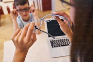 Close up de mãos femininas segurando óculos enquanto criança do sexo masculino sentado à mesa com laptop moderno