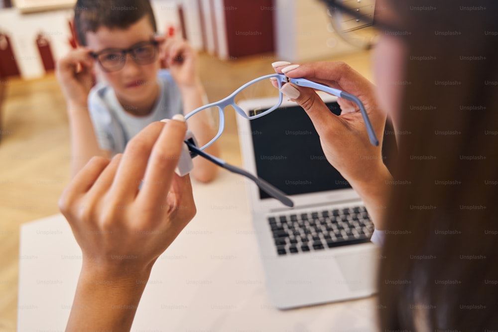 Nahaufnahme von weiblichen Händen, die eine Brille halten, während männliches Kind mit modernem Laptop am Tisch sitzt