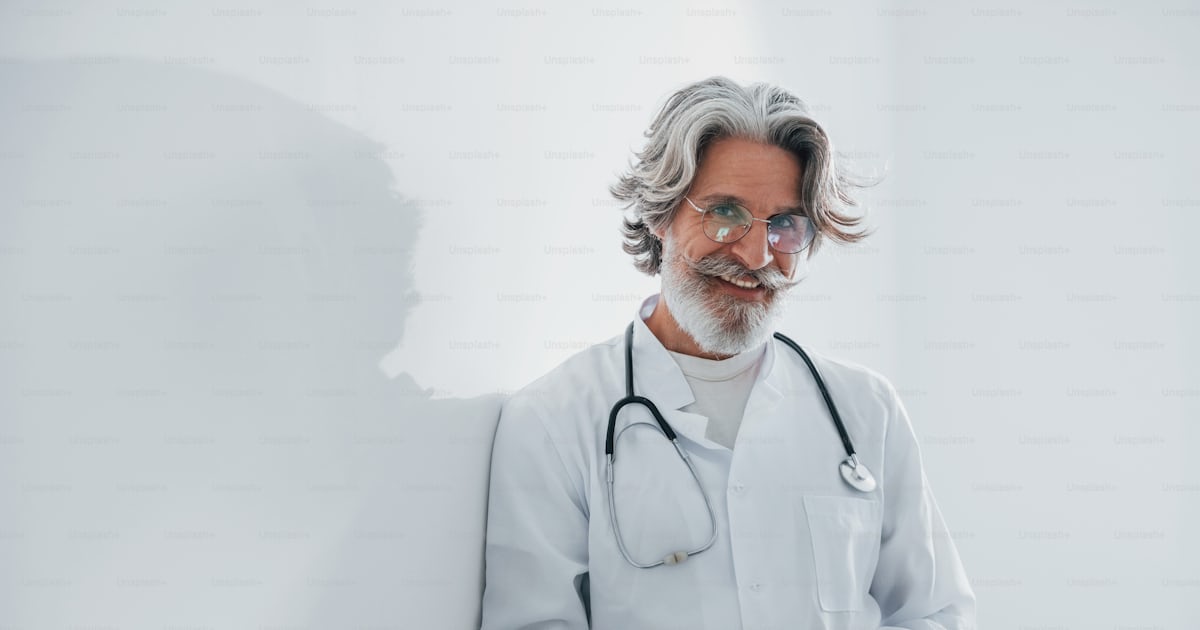 Foto zum Thema Oberarzt mit grauen Haaren und Bart im weißen Kittel ist