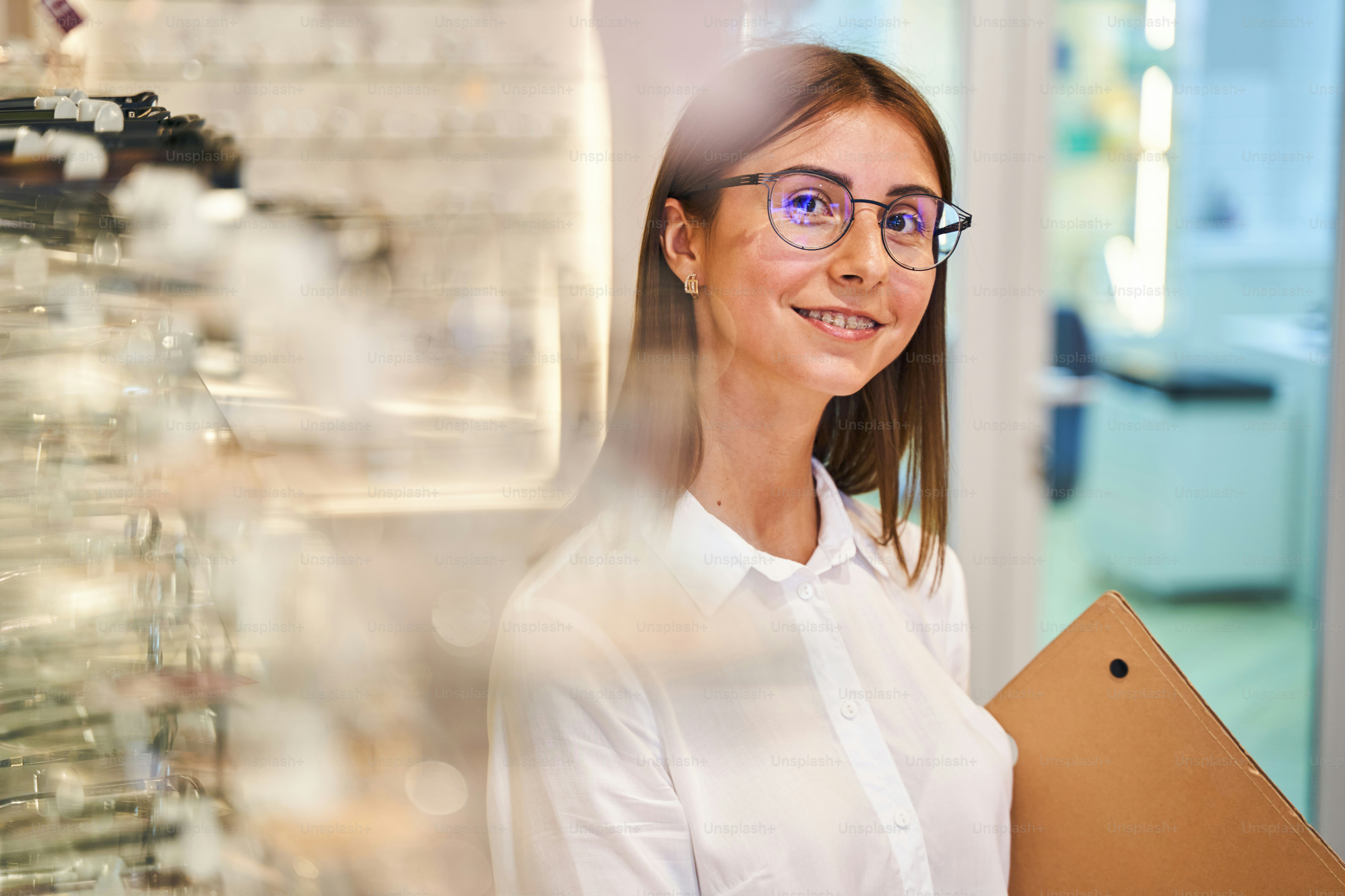 Charming young woman holding folder and smiling while standing by showcase in glasses store