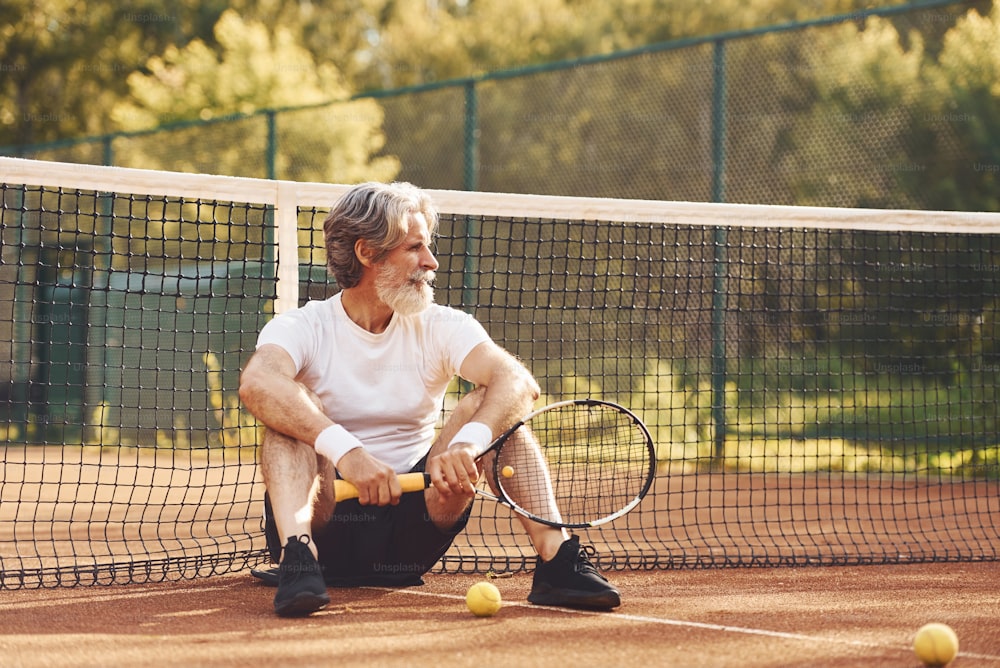 Sedersi e fare una pausa. Uomo alla moda senior in camicia bianca e pantaloncini sportivi neri sul campo da tennis.