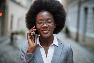 Portrait of smiling african lady in stylish suit and eyeglasses using modern smartphone for conversation outdoors. Concept of business, people and technology.